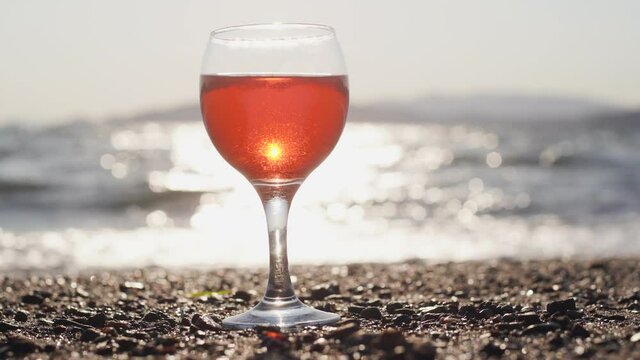 Close-up view 4k stock video footage of one glass of pink wine standing on beach isolated on sunny sunset blue sky and blurry sea water background