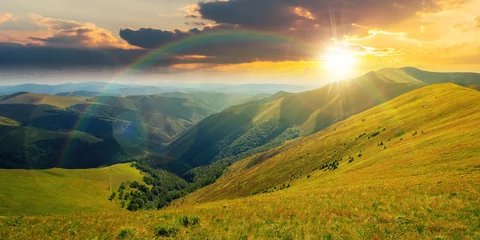 Poster mountain landscape in summer at sunset. grassy meadows on the hills rolling in to the distant peak beneath a rainbow in evening light © Pellinni