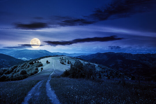 road through meadow in mountains at night. beautiful rural landscape of carpathians in full moon light. wonderful summer weather with fluffy clouds on the sky