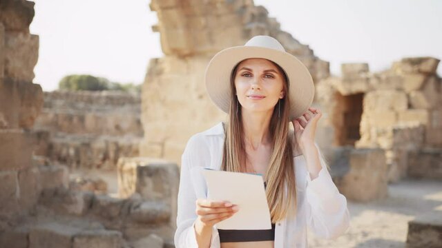 Woman in white hat and casual clothes walking among ruined ancient temple with map in hands. Archeologist discovering abandoned historical places. Archeologist examining old historical buildings