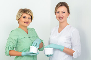 Fototapeta premium Two cosmeticians with jars of wax for depilation smiling against a white background. Natural product for hair removal. Copy space