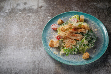 Serving a dish from the restaurant menu. Fresh Caesar salad of chicken with croutons, tomatoes and herbs on a blue plate against a gray stone table, a delicious appetizer