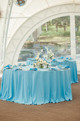 Wedding tables for guests. Round tables with a blue tablecloth. Decorated with flowers, plates, glasses and more