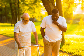Obraz premium Caregiver is teaching old man to walk with walker. Professional nurse and patient walking outdoor at sunset. Assistance, rehabilitation and health care.