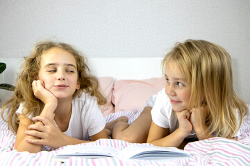 two cute sisters are lying on the bed and reading a funny book, smiling and laughing, family evening, one looking at the other, children's development through independent reading