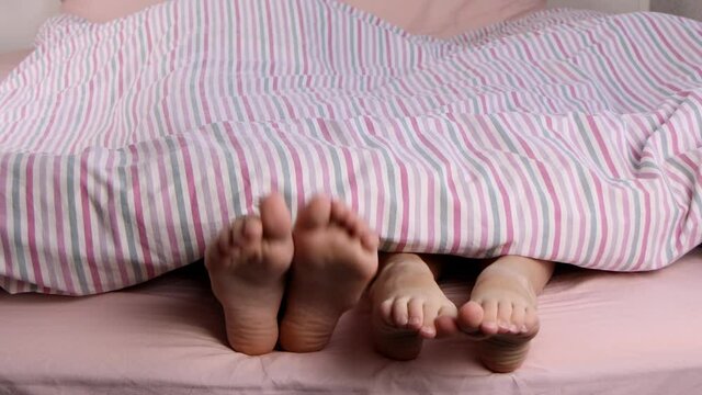 Children s legs stick out from under the striped blanket. Little fingers and heels. Baby feet. Children lie in bed barefoot and move their feet. Cozy home morning, awakening.