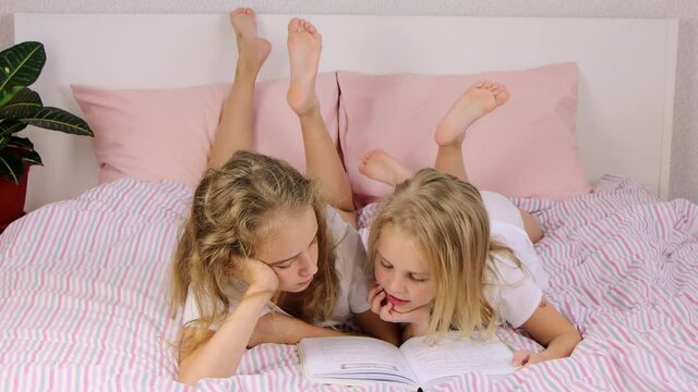 two cute little girls sisters lie on the bed and read a funny book, smile and laugh, family evening, bedtime, reading by children