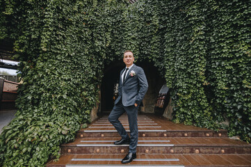 Fototapeta premium Stylish groom in a gray suit stands on the steps in nature, against the backdrop of green ivy foliage. Wedding portrait.