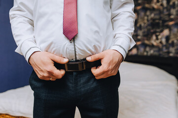 A man, a groom, a businessman in a white shirt and a red tie puts a leather belt on black trousers, going to a wedding, wedding in the morning.