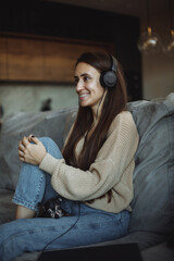 A smiling young brunette girl with long hair in a beige jumper and blue jeans sits at home on a gray sofa and listens to music on headphones. At the feet of a dog breed toy terrier