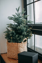 Wicker basket with a Christmas tree decorated with a garland at home on the windowsill, next to it is a wrapped black gift