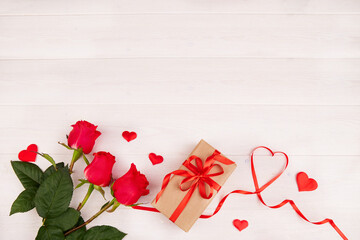 A bouquet of red roses, a box in a craft paper package with a red ribbon and a heart-shaped ribbon on a white wooden background. Valentine's Day, Mother's Day, Birthday. Top view, copy space.