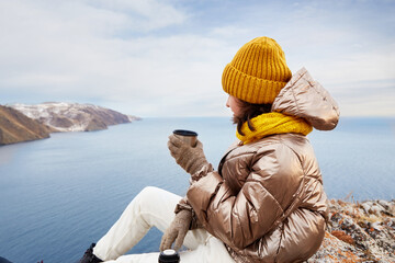 A young girl is a traveler at a rest stop. Admiring the beautiful winter landscape, drinking hot tea from a thermos. Freedom, the concept of travel, active recreation. 
