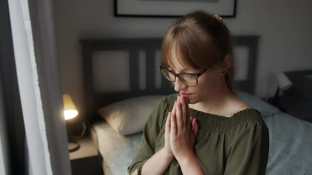 Young Religious Woman Saying Morning Prayer WIth Folded Hands, Sitting on Bed by Window, Close Up