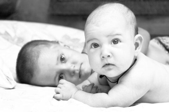Caucasian toddler boy and his baby sister are lying on bed and looking at camera. Close up portrait. Black and White horizontal shot. Sibling rivalry concept