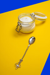thick greek yogurt in glass jar and vintage dessert spoon on yellow and blue background