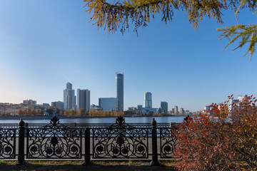City autumn landscape made in Yekaterinburg (Russia) on a sunny day. River embankment with tall modern buildings. In the foreground there is an openwork forged trellis and autumn trees and bushes 