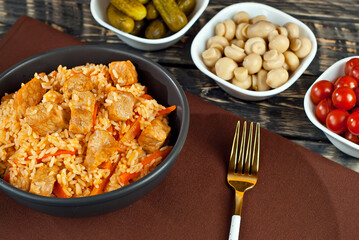 Pilaf with meat and carrots in a black bowl. Marinated mushrooms, tomato and cucumber on a black background. Food on a shabby table. Bowl on an old wooden board.