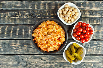 Pilaf with meat and carrots in a black bowl. Marinated mushrooms, tomato and cucumber on a black background. Food on a shabby table. Bowl on an old wooden board.