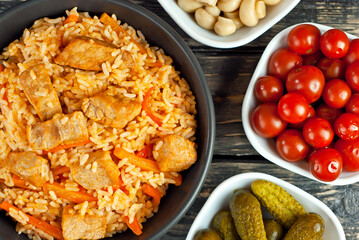 Pilaf with meat and carrots in a black bowl. Marinated mushrooms, tomato and cucumber on a black background. Food on a shabby table. Bowl on an old wooden board.