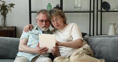 Happy aged wife sitting on cozy sofa hugging mature hoary husband scrolling web pages on digital touchpad in his hands. Smiling relaxed senior couple using tablet discussing news video blog having fun - Powered by Adobe