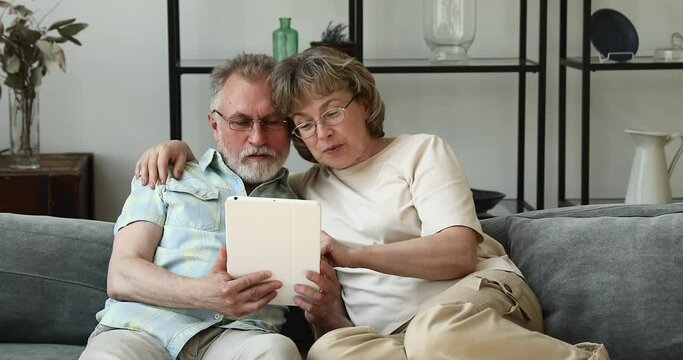 Happy aged wife sitting on cozy sofa hugging mature hoary husband scrolling web pages on digital touchpad in his hands. Smiling relaxed senior couple using tablet discussing news video blog having fun