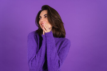 Fototapeta premium Young woman in a purple soft cozy sweater on the background is cute, in high spirits, laughs shyly, covering her mouth with her hands