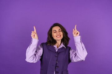 Young woman in a shirt and a vest on a purple background smiles cheerfully in high spirits, points up with a finger to the empty space