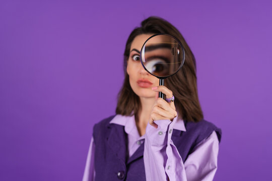Young woman in shirt and vest on purple background having fun with magnifying glass in hand
