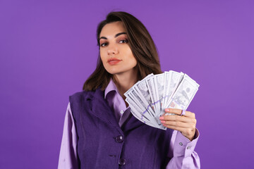 Young woman in a shirt and vest on a purple background with a bundle of money dollars in her hand smiles confidently