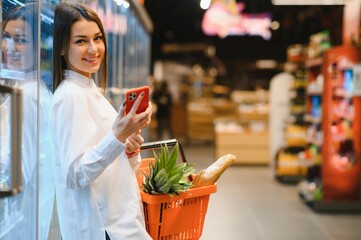 Woman doing grocery shopping at the supermarket, she is leaning on the shopping cart and connecting with her phone, apps and retail concept