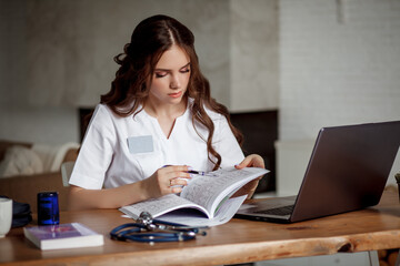 A beautiful nurse in a white medical suit sits in front of a laptop at her desk. Medical concept.