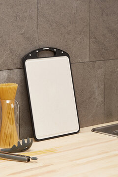 Shot of a cutting board with black inserts at the edges. It stands next to a gray wall. A glass jar filled with spaghetti and a pasta spoon are next to the cutting board. All of the items are on a woo