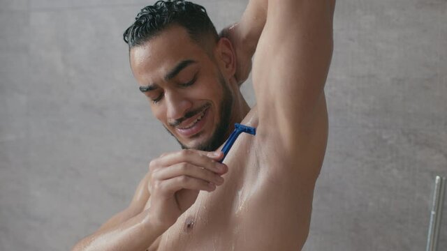 Hygienic procedures. Young nude happy middle eastern man shaving his armpit while taking morning shower at home