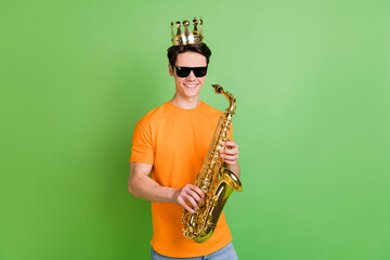 Photo of cheerful young happy man wear crown good mood hold saxophone isolated on green color...