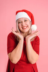 An adult woman in a Santa hat supports her chin with her hands standing on a pink background, vertical photo