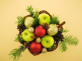  New Year's  arrangement of fir branches,  green apples, Christmas tree red and gold balls. Winter decorations  for the interior. Fruit basket  on yellow  background. 