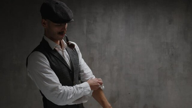 Young dangerous gangster man dressed in vintage suit, cap and smoking pipe, warming up, rolls up his sleeves, prepares for fight, kneads his fists and neck in front of concrete wall