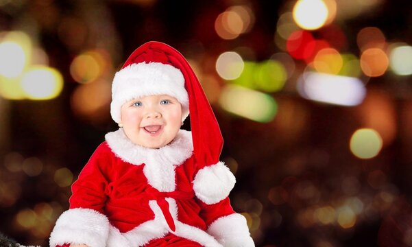 Cute little baby wearing Santa hat against blurred lights on dark background. Christmas celebration
