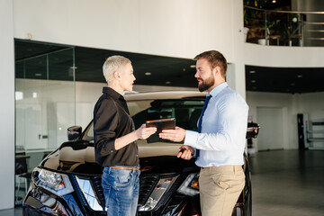 Concept of expensive purchase. Smiling friendly male car seller with tablet in hands talking about car specifications to a woman customer in black shirt who wants to buy a car. Car salon interior.