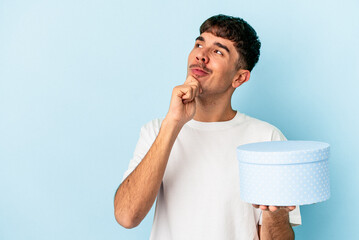 Young mixed race man holding a box present isolated on blue background looking sideways with...