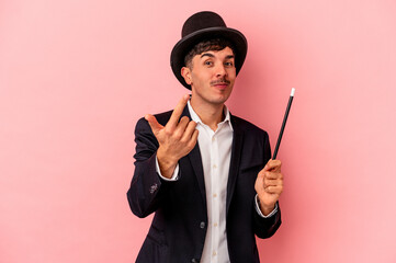 Young caucasian wizard man holding a wand isolated on pink background pointing with finger at you as if inviting come closer.