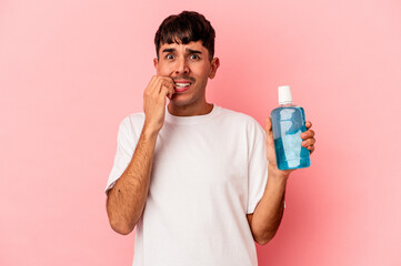 Young mixed race man holding mouthwash isolated on pink background biting fingernails, nervous and very anxious.