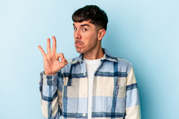 Young mixed race man isolated on blue background winks an eye and holds an okay gesture with hand.