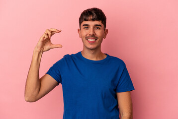 Young mixed race man isolated on pink background holding something little with forefingers, smiling and confident.