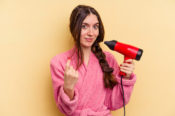 Young woman holding a hairdryer isolated on yellow background pointing with finger at you as if inviting come closer.
