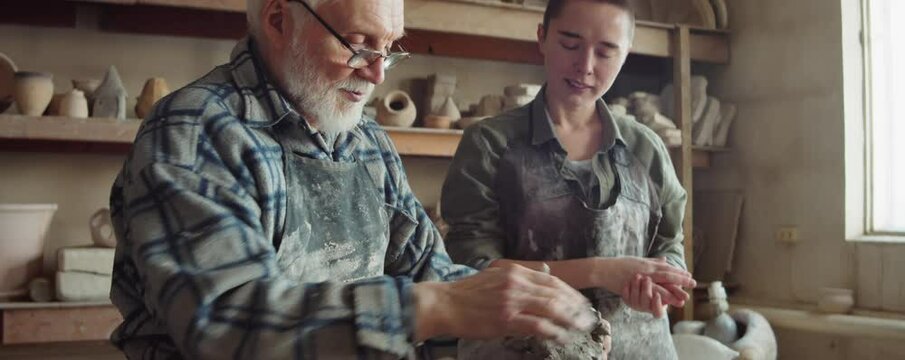 Elderly sculptor forming clay head and giving lesson to young female apprentice in pottery studio