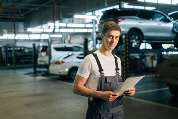 Medium shot portrait of smiling handsome young mechanic male wearing uniform holding clipboard standing in auto repair shop garage, with vehicle background, looking at camera