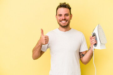 Young caucasian man holding a iron isolated on yellow background smiling and raising thumb up