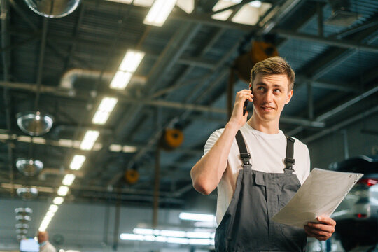 Low-angle view of professional handsome young mechanic male wearing uniform holding clipboard and talking on mobile phone, standing in auto repair garage, with vehicle background, looking at camera